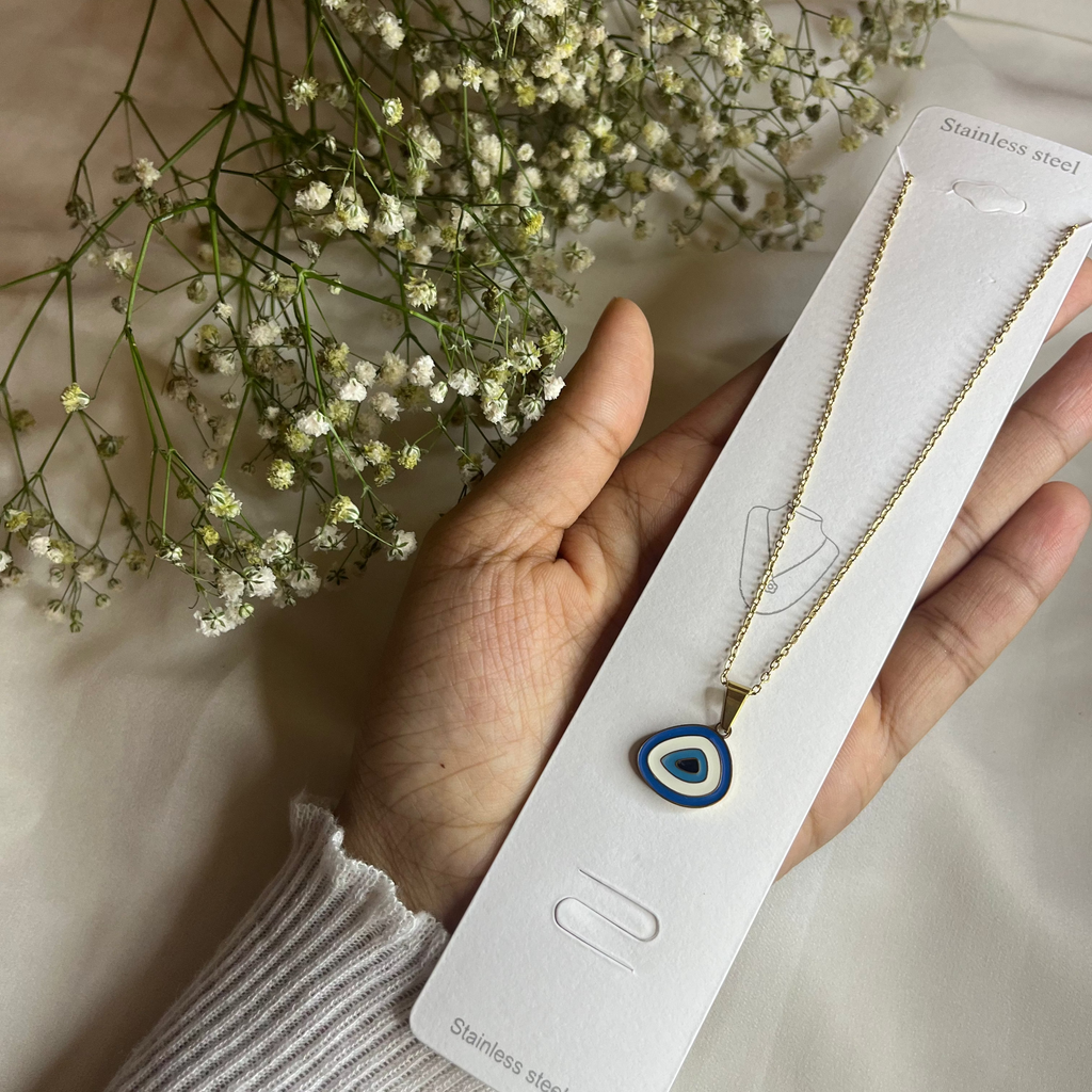 Hand holding a necklace with an evil eye pendant on a white card, with flowers in the background.
