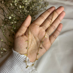 Hand wearing a delicate gold necklace with small beads, surrounded by dried flowers on a neutral background.